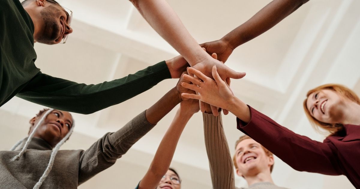 Fünf motivierte Mitarbeitende stehen im Kreis und legen als Team die Hände zusammen – fotografiert von unten