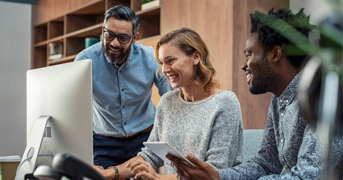 Drei lächelnde Mitarbeitende sitzen gemeinsam vor einem Computer und schauen auf den Bildschirm. Eine Person hält ein Tablet in der Hand.